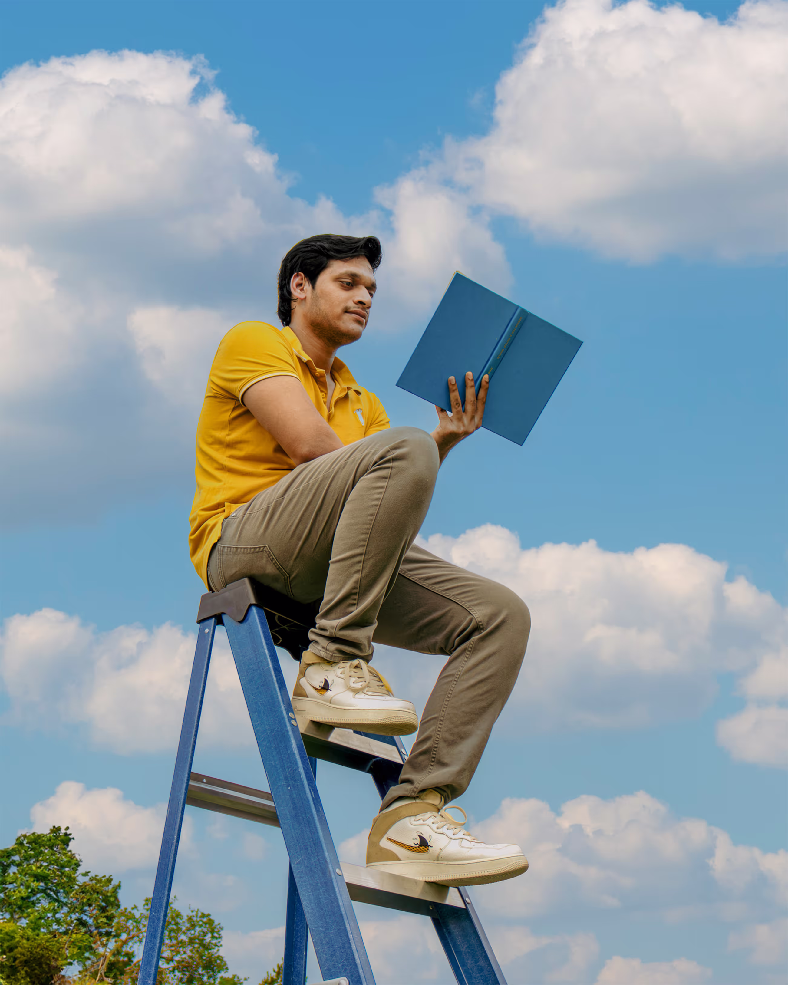 Person sitting on a ladder in yellow shirt reading a book with cloudy background.